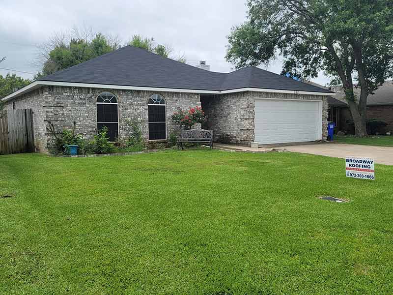 Small home with new dark gray shingles installed by Broadway Roofing with their yard sign in front